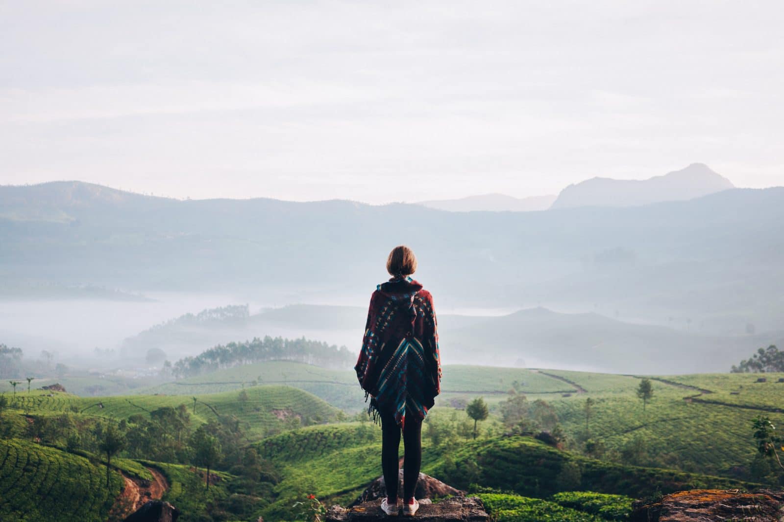 KI-basierte Bildbeschreibung | Eine Person steht auf einem Felsen und blickt auf grüne Hügel und Felder mit Nebel und Bergen im Hintergrund unter einem bewölkten Himmel.